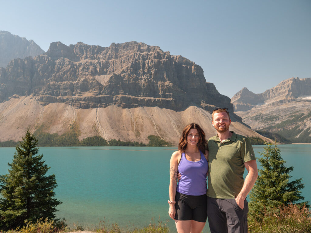 a man and woman standing next to a body of water with mountains in the background
