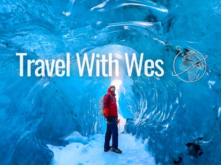 Man in an orange coat standing inside a blue glacier ice cave, Travel with Wes logo on image