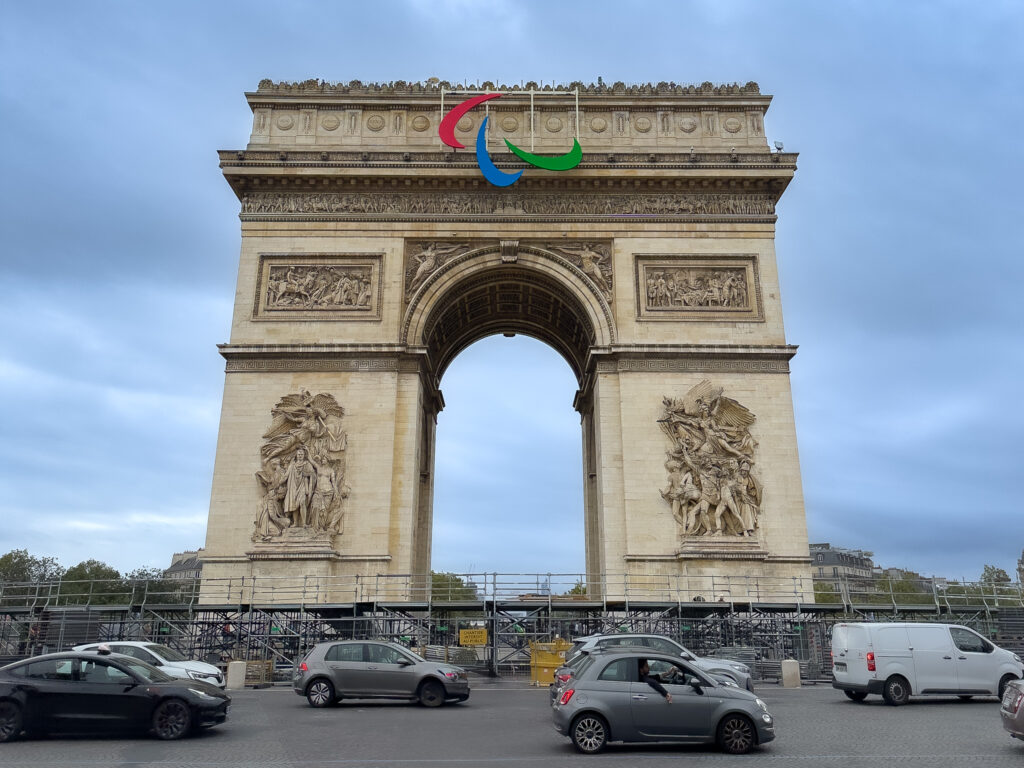 a large stone arch with cars parked in front of it with Arc de Triomphe in the background