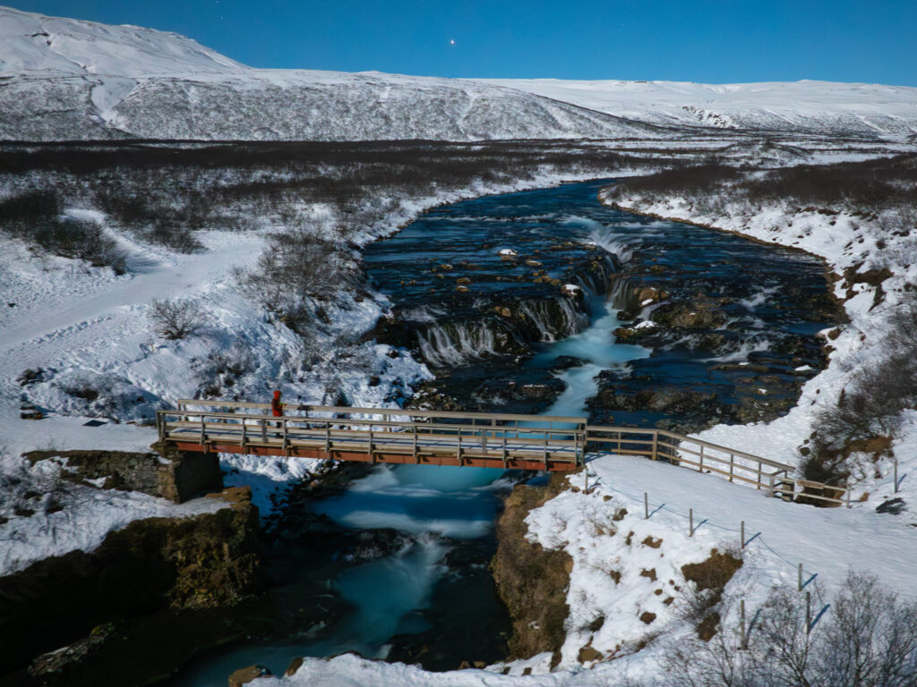 a bridge over a river with a waterfall and snow covered hills