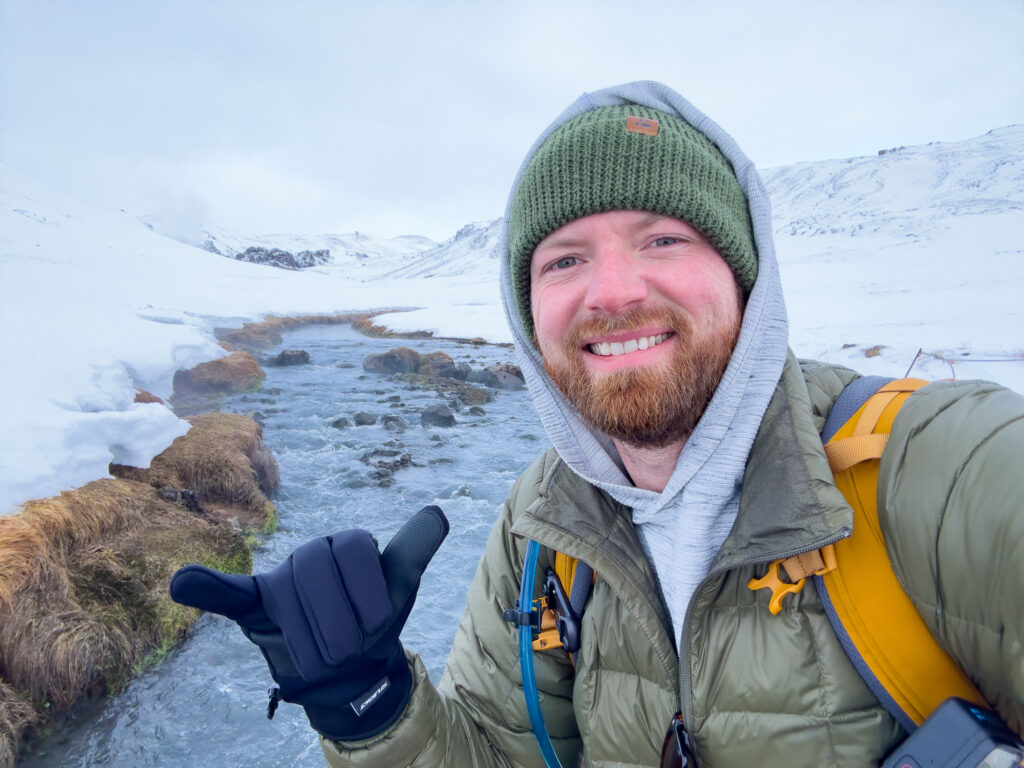 a man in a hat and a green beanie pointing his finger up in front of a river